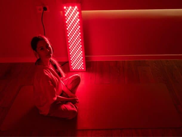 A woman sitting on a yoga mat beside a glowing red light therapy panel for wellness and recovery